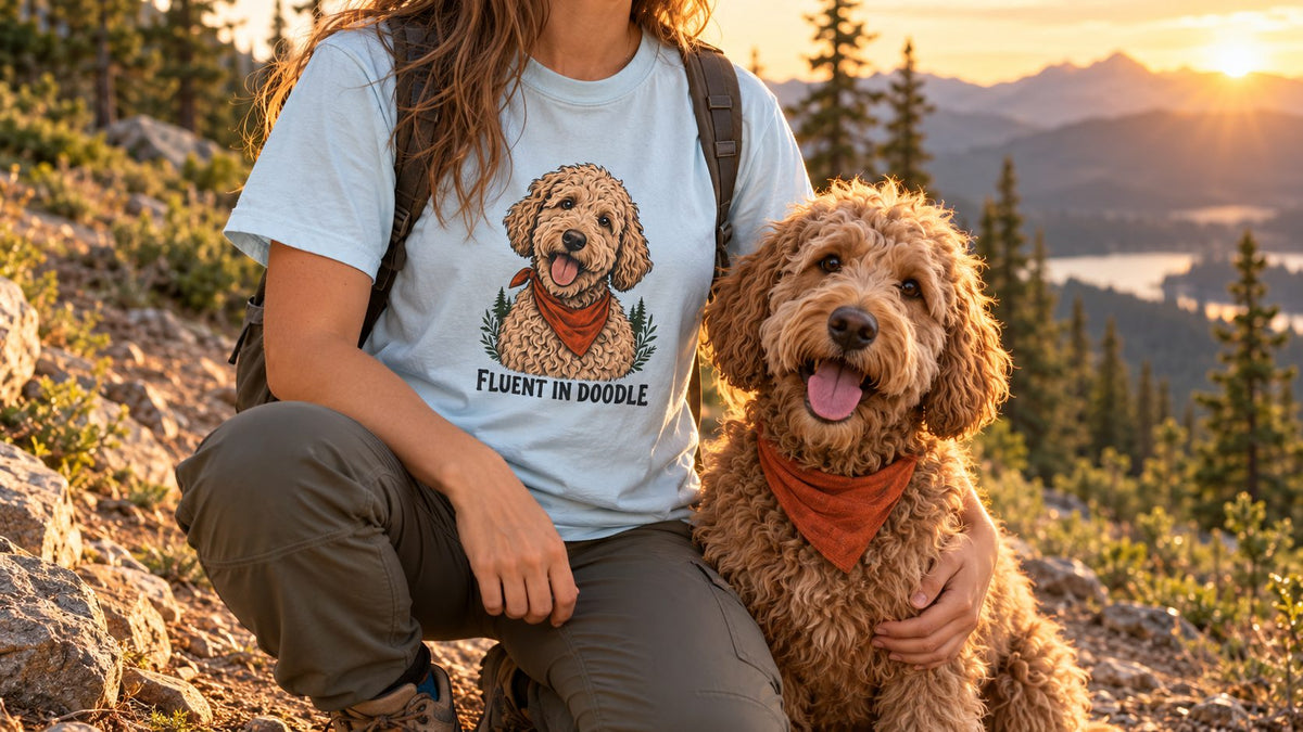 Hiker in a light blue Fluent in Doodle tee kneeling on a mountain trail beside a happy doodle in an orange bandana