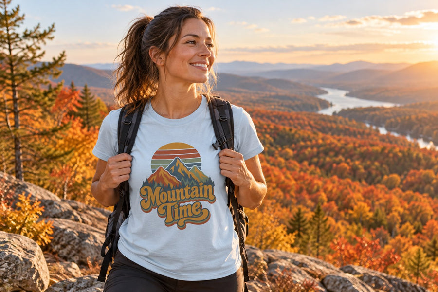 Smiling hiker in a light blue Mountain Time tee at a rocky overlook above orange and gold fall forest and a winding river