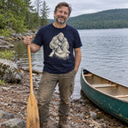 Man wearing the Bigfoot Canoe T-Shirt by a lake with a canoe