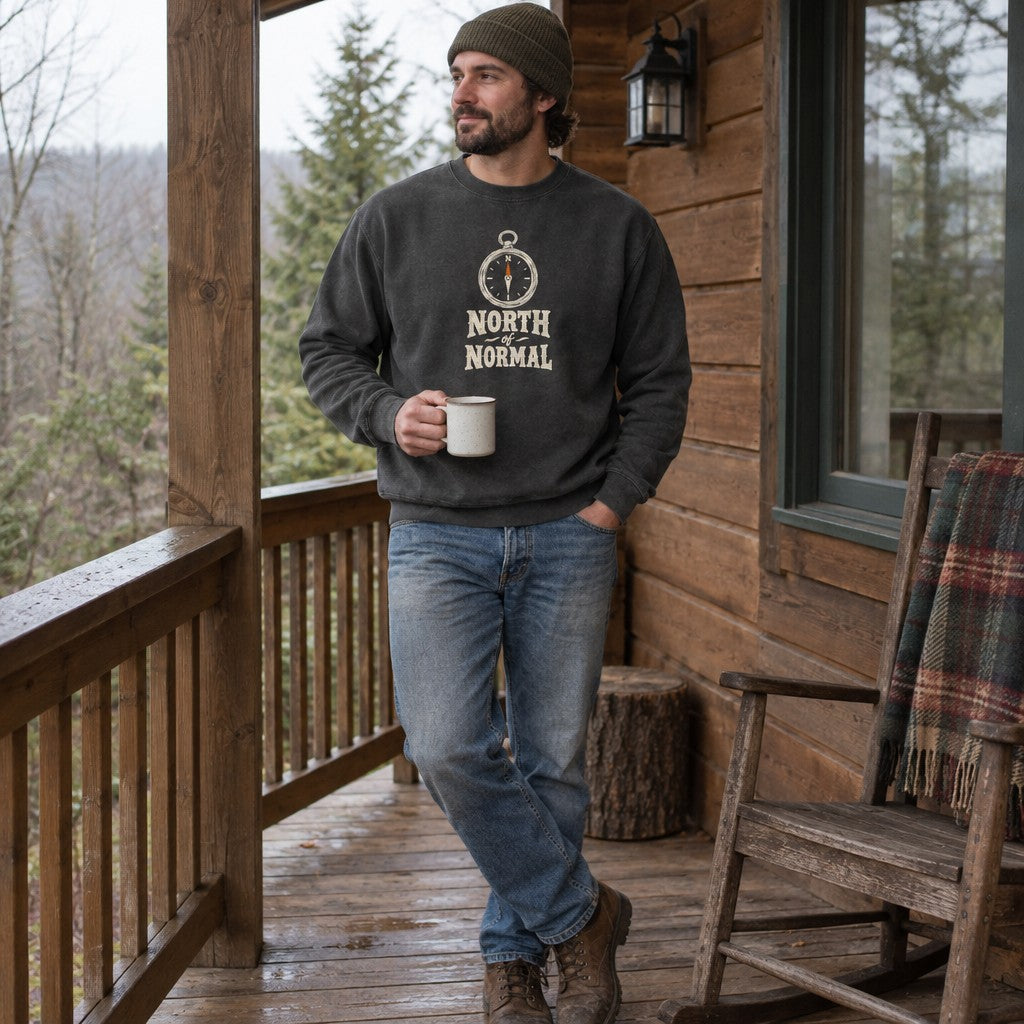 Man wearing the North of Normal crewneck sweatshirt on a cabin porch with a mug