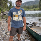 Man wearing the Follow the River T-Shirt on a riverbank next to a canoe