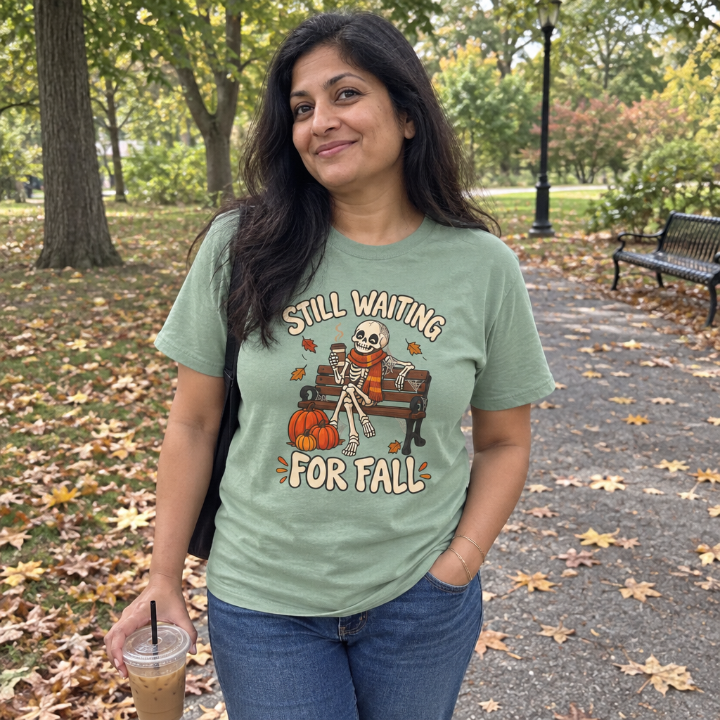 Woman wearing the sage green Still Waiting for Fall t-shirt on a leafy park path with iced coffee