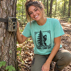 Woman wearing the Trail Cam Shy Bigfoot t-shirt next to a trail camera