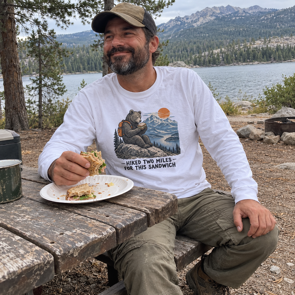 Man wearing the white Hiked Two Miles for This Sandwich long sleeve tee at a lakeside campground