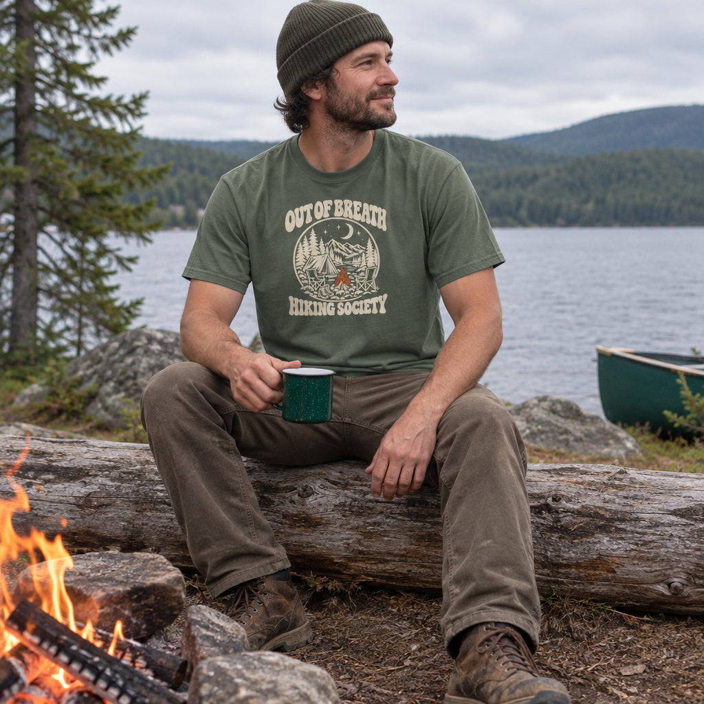 Man wearing the Out of Breath Hiking Society Campfire Night T-Shirt at a lakeside campfire with a mug