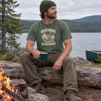 Man wearing the Out of Breath Hiking Society Campfire Night T-Shirt at a lakeside campfire with a mug