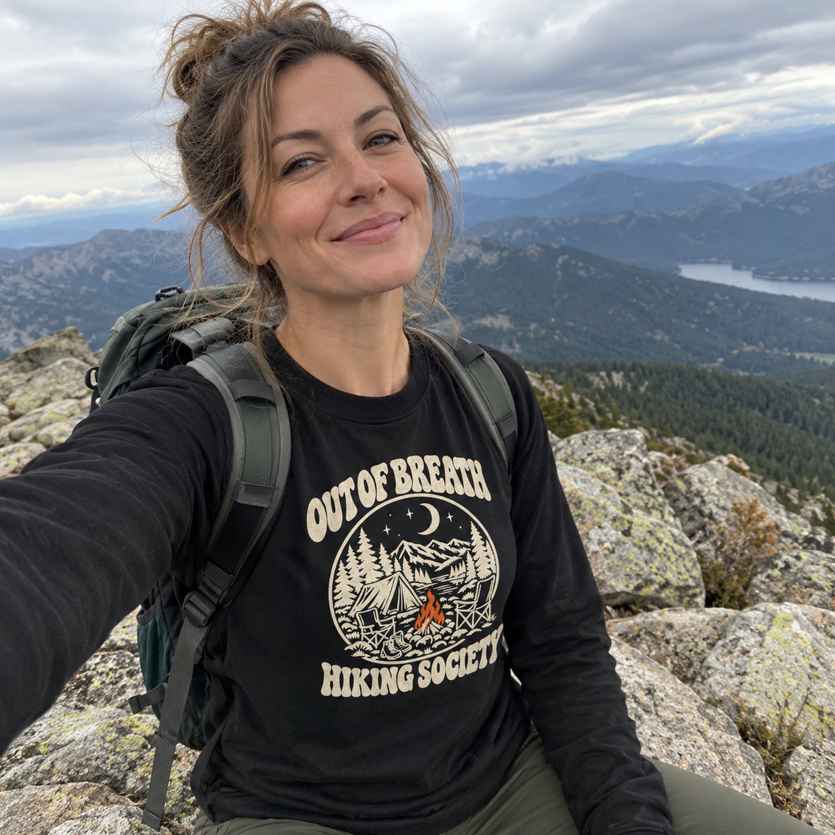 Woman wearing the Out of Breath Hiking Society Campfire Night Long Sleeve Shirt taking a selfie on a mountain summit with a backpack