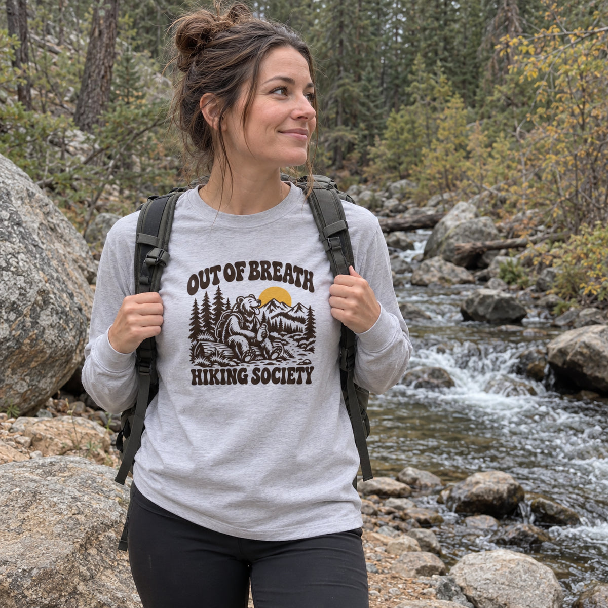 Woman wearing the Out of Breath Hiking Society Bear Long Sleeve Shirt by a forest creek with a backpack