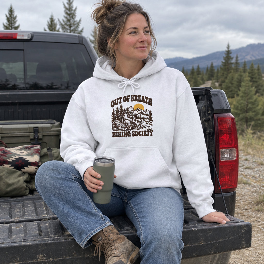 Woman wearing the Out of Breath Hiking Society Bear Hoodie on a truck tailgate with a tumbler
