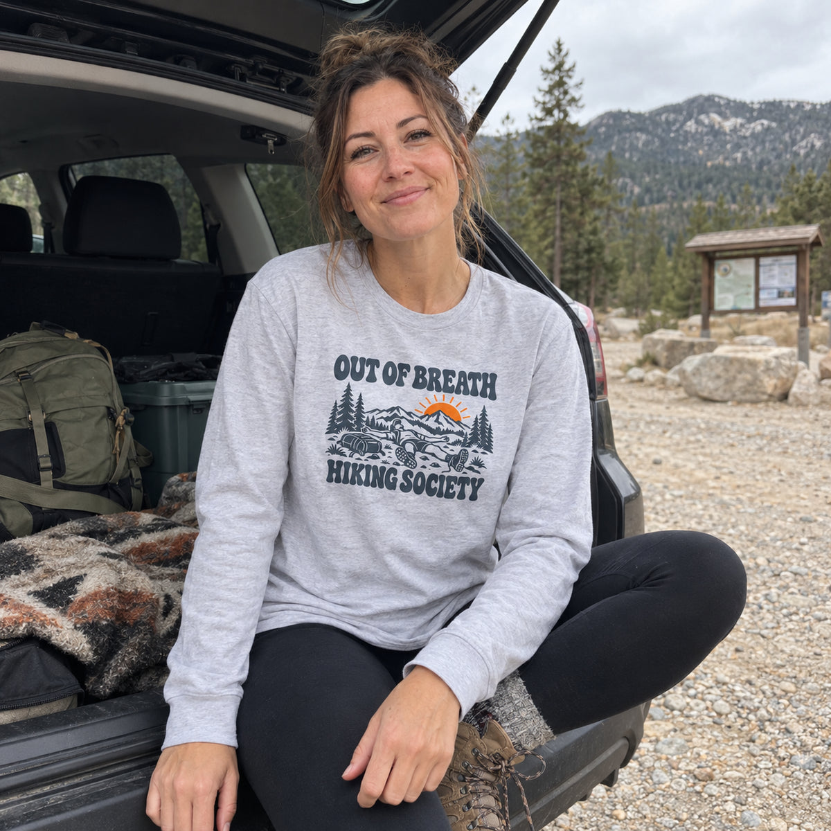 Woman wearing the Out of Breath Hiking Society Flat Out Long Sleeve Shirt at the open back of a car at a trailhead