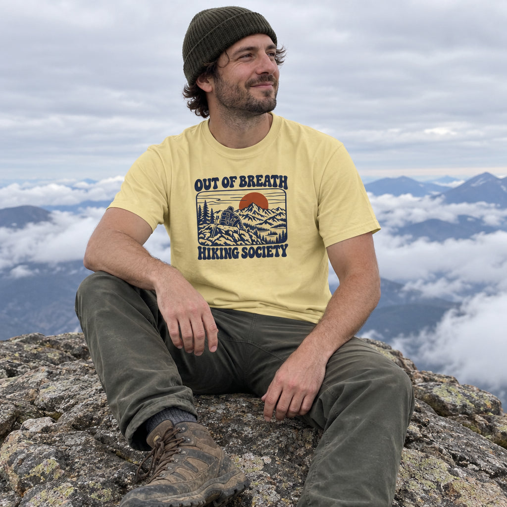 Man wearing the Out of Breath Hiking Society Summit Rest T-Shirt sitting on a mountain summit above the clouds
